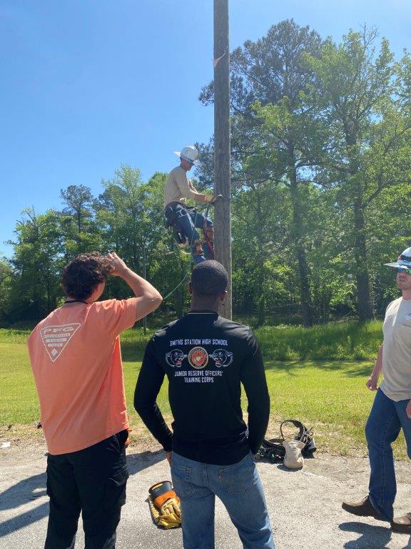 Lineman Mitchell Greene, on pole, and Lineman Preston Hancock, right, walk students through a pole-climbing demonstration at the lineworker career day in Phenix City. (Brooke Goff / Alabama News Center)